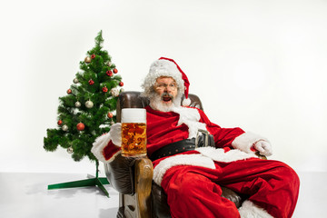 Santa Claus drinking beer near the Christmas tree, congratulating, looks drunk and happy. Caucasian male model in traditional costume. New Year 2020, gifts, holidays, winter mood. Copyspace for your