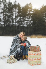 Mother and baby sitting on wooden sledge in winter forest and drinking hot tea. Christmas gifts presents on the snow. Winter snowy forest on the background. Christmas vacation.