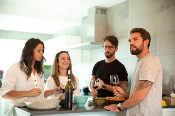 Team of friends chatting over glass of wine in kitchen. Young men and women in casual meeting indoors. Friendship concept