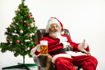 Santa Claus drinking beer near the Christmas tree, congratulating, looks drunk and happy. Caucasian male model in traditional costume. New Year 2020, gifts, holidays, winter mood. Copyspace for your