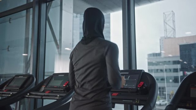 Athletic Muslim Sports Woman Wearing Hijab and Sportswear Running on a Treadmill. Energetic Fit Female Athlete Training in the Gym Alone. Urban Business District Window View. Back View Zoom in