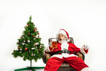 Santa Claus drinking beer near the Christmas tree, congratulating, looks drunk and happy. Caucasian male model in traditional costume. New Year 2020, gifts, holidays, winter mood. Copyspace for your