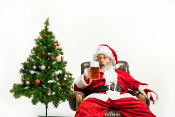 Santa Claus drinking beer near the Christmas tree, congratulating, looks drunk and happy. Caucasian male model in traditional costume. New Year 2020, gifts, holidays, winter mood. Copyspace for your