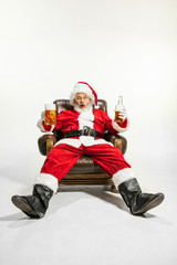 Santa Claus drinking beer sitting on armchair, congratulating, looks drunk and happy. Caucasian male model in traditional costume. New Year 2020, gifts, holidays, winter mood. Copyspace for your ad.