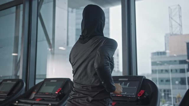 Athletic Muslim Sports Woman Wearing Hijab and Sportswear Running on a Treadmill. Energetic Fit Female Athlete Training in the Gym Alone. Urban Business District Window View. Back View Zoom Out