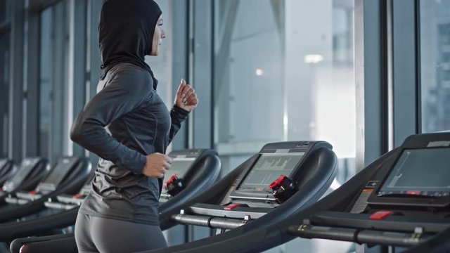 Athletic Muslim Sports Woman Wearing Hijab and Sportswear Running on a Treadmill. Energetic Fit Female Athlete Training in the Gym Alone. Urban Business District Window View. Side View Portrait