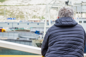 Dover, England, 15/05/2019 Man looking over the bannister of a P&O ferry at the coast of dover with a coat grey hair old glasses and beard vaping on a ship boat with coat on cold weather