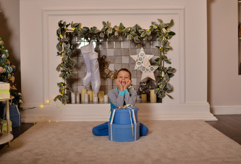 A white child sits with a gift near a large decorated spruce tree in a beautiful fireplace interior. Large decorated Christmas tree in blue and light colors