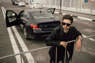 a man in sunglasses and a leather jacket stands near a sports car