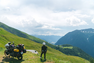 Biker woman wearing protective turtle jacket. standing near his tourer motorcycle on the green grass. Looking into distance. Alpine mountains.