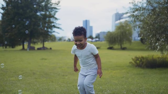 African baby boy runs around the park and catches air bubbles. Happy child outdoors.
