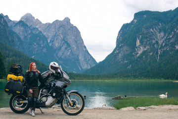 Girl motobiker in protective turtle jacket stands on the beach with touring motorcycle. Extreme travel vacation, motorcyclist adventure lifestyle. Toblacher See, (Lago di Dobbiaco) Italy. Copy space