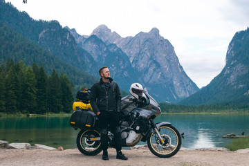 Fototapeta na wymiar Handsome man in the center of the composition with motorcycle on beach. Alpine mountains and lake on background. Biker lifestyle, world traveler. Toblacher See, (Lago di Dobbiaco) Italy.