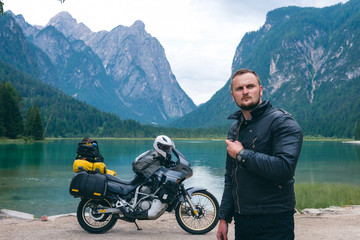 Portrait of handsome man motorcyclist with touring motorcycle on beach. Alpine mountains and lake on background. Biker lifestyle, world traveler. Toblacher See, (Lago di Dobbiaco) Italy Ready to ride