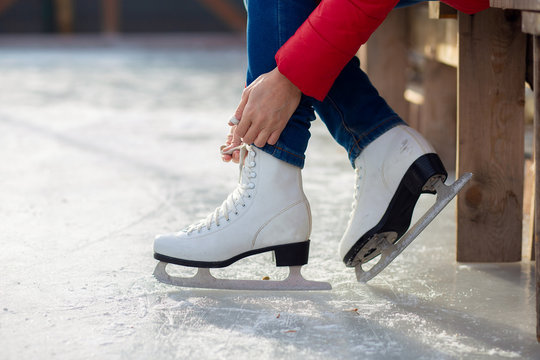 A girl in a red jacket is tying shoelaces on figured white skates on an ice rink