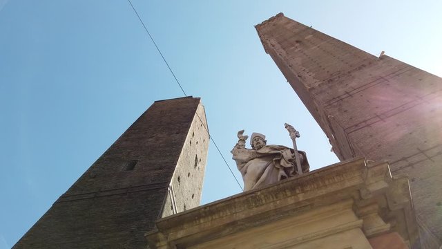 Perspective view of Due Torri or two towers: Torre degli Asinelli and Torre Garisenda, a symbol of Bologna city, Italy, in historic center of the city in the blue sky with saint San Petronio statue.