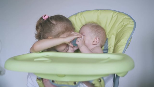 cute girl helps junior brother sitting in soft green highchair drink water from bottle in light room closeup