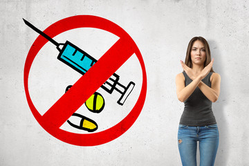 Serious young girl with hands crossed, standing against wall with picture of syringe and some meds...
