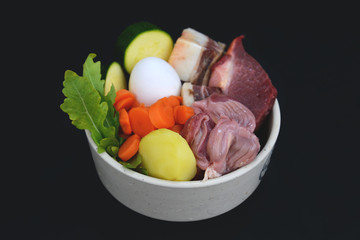 Top view of dog bowl filled with mixture of biologically appropriate raw food containing meat chunks, vegetables, egg on dark black background