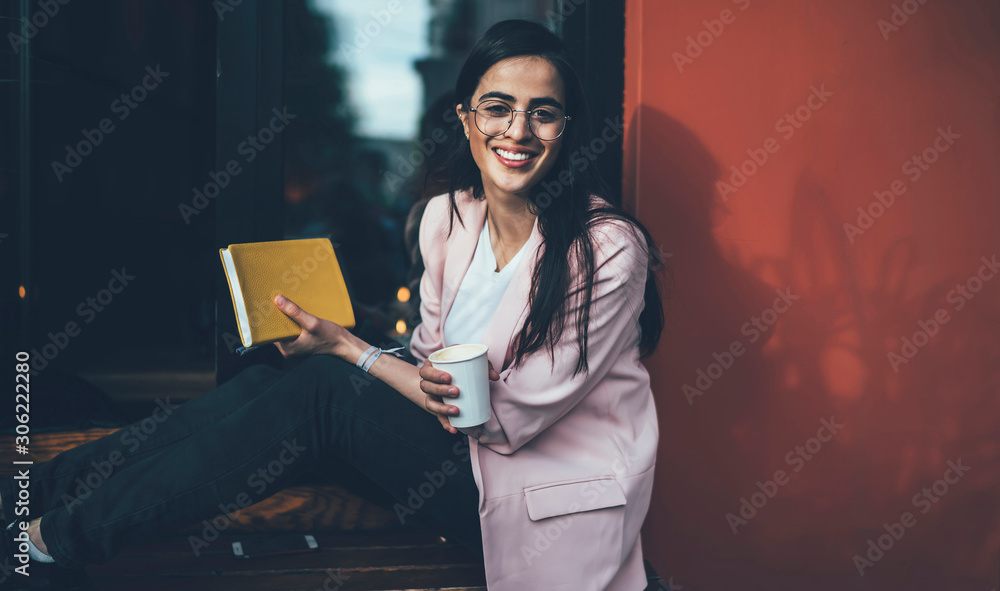 Poster portrait of happy female author of web blog for students sitting near coffee shop with caffeine beve