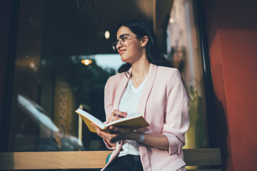 Positive female writer sitting outdoors and creating new charter of best seller thoughtful looking away and thinking on positive idea for making notes in knowledge textbook, notepad for education