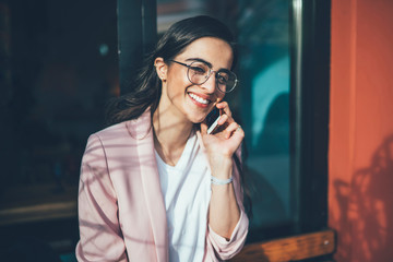 Smiling young woman in spectacles talking on phone with friend satisfied with mobile connection, happy hipster girl getting good news during conversation on smartphone spending time outdoors