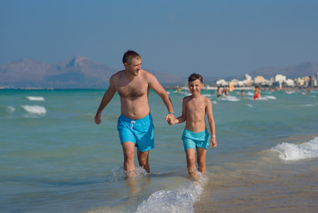 Dad and son are running in the water along the sea shore. They are holding hands and moving towards the camera.