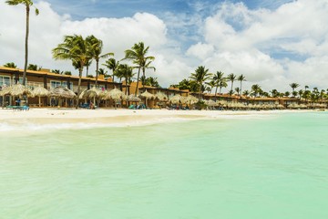 Beautiful view of white sand Aruba beach. Blue sunbeds under sun umbrellas on turquoise water and blue sky with white clouds background. Eagle Beach of Aruba Island.