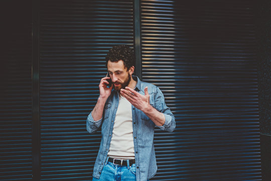 Angry bearded young man gesturing hands while solving problem during phone conversation on smartphone.Annoyed hipster blogger has quarrel while calling on mobile phone standing outdoors against wall