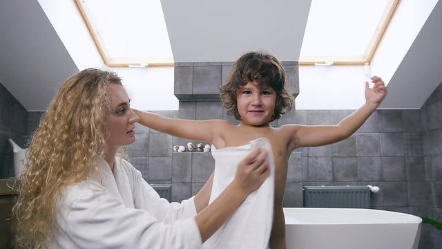 Smiling handsome boy showing arm gestures how he is strong while his cute blond mother wiping the body with towel