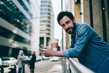 Portrait of positive bearded hipster guy having coffee break outdoors on urban settings using...