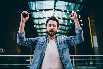 Portrait of emotional hipster guy in casual wear raising hands standing outdoors celebrating victory raising hands and screaming, overjoyed cheerful male expressing success and happiness win.