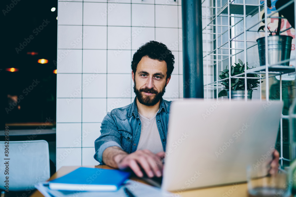 Poster portrait of bearded hipster guy looking at camera working at laptop computer in office,skilled busin