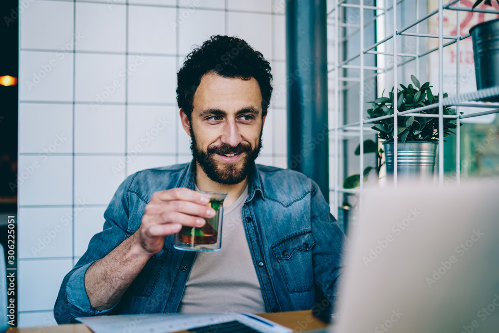 Poster bearded young man dressed in denim wear and drinking tea while watching tutorial online on modern la