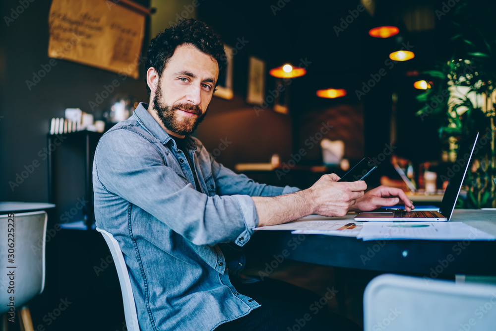 Poster portrait of handsome bearded hipster guy holding smartphone in hand while sharing media on laptop co