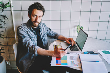 Portrait of bearded young man holding smartphone in hand and looking at camera during remote work at laptop in coworking.Hipster guy chatting on phone sitting at desktop with paper diagrams