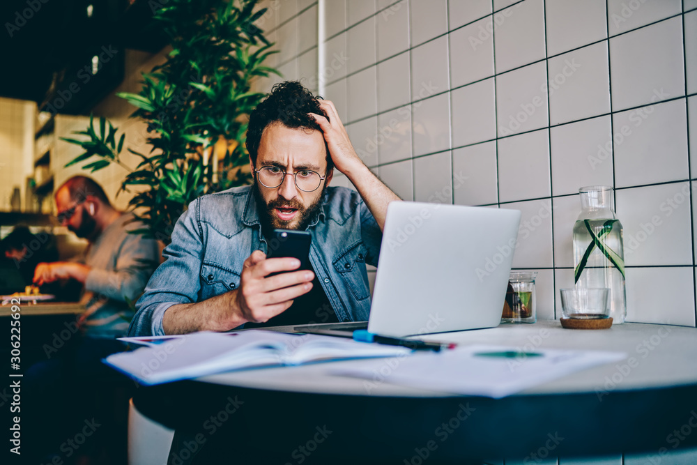 Poster emotional guy in eyewear shocked while reading mail notification on mobile phone, expressed disappoi