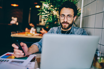 Portrait of bearded young man freelancer checking email on mobile phone while looking at camera and listening audio player downloaded on laptop in earphones sitting at desktop in coworking space