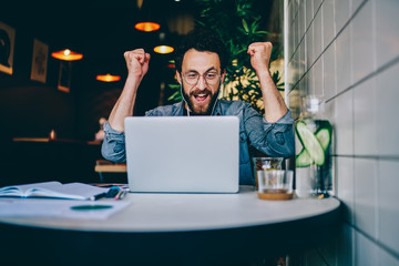 Overjoyed hipster guy in earphones cheers for team watching match on laptop computer during free...