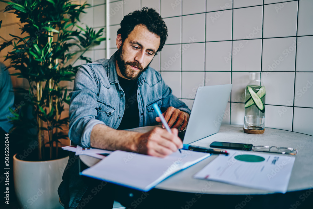 Poster pensive bearded young man studying at laptop computer and writing down notes from internet website o