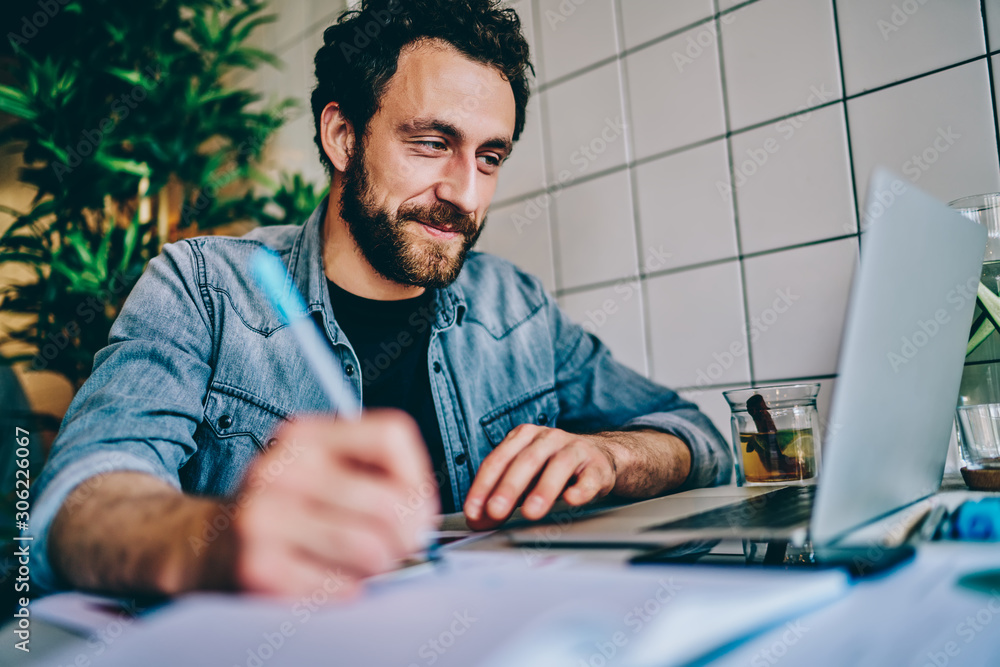 Poster cheerful bearded young man watching webinar online on website and writing down text notes for studyi