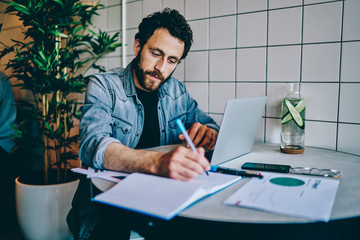 Pensive bearded young man studying at laptop computer and writing down notes from internet website...