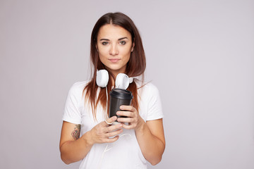 Serious mixed race woman enjoys hot drink in cold weather, holds black disposable cup with coffee, dressed in blank t-shirt,headphones, look directly at camera, poses indoor, needs morning refreshment