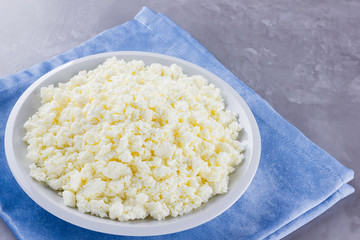 Cottage cheese in a plate. Fresh cottage cheese in white plate on a gray background. Soft cheese on a blue napkin. Top view. Copy space