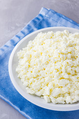 Cottage cheese in a plate. Fresh cottage cheese in white plate on a gray background. Soft cheese on a blue napkin. Top view. Copy space