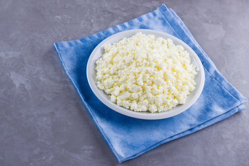 Cottage cheese in a plate. Fresh cottage cheese in white plate on a gray background. Soft cheese on a blue napkin. Top view. Copy space