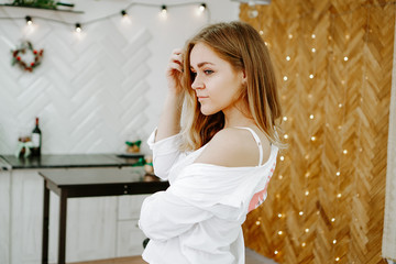 A girl in a long white shirt stands in the kitchen. The white kitchen is decorated with Christmas decorations and garlands