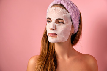Portrait of a girl with a bandage on her head, a fabric mask on her face and bare shoulders on a pink background.