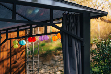 Little colorful lampions hanging on a pavilion in a garden on a sunny summer day. Cozy Chairs and...