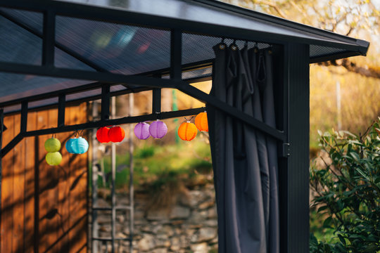 Little colorful lampions hanging on a pavilion in a garden on a sunny summer day. Cozy Chairs and Table standing underneath it.